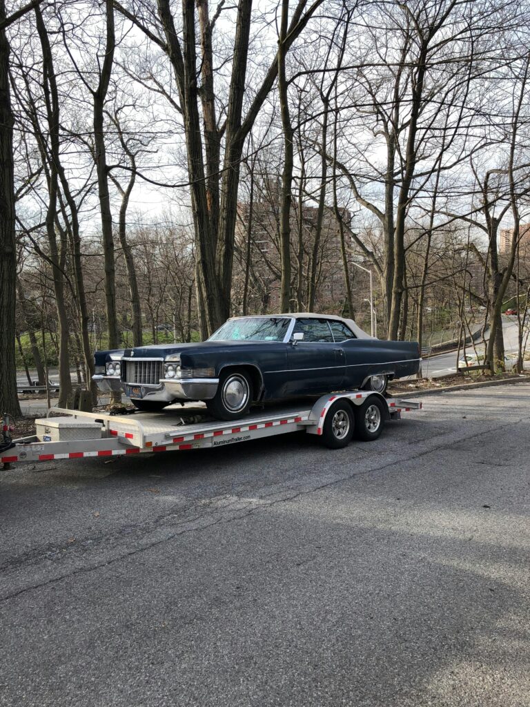 Classic blue car on a trailer amidst leafless trees on a street.